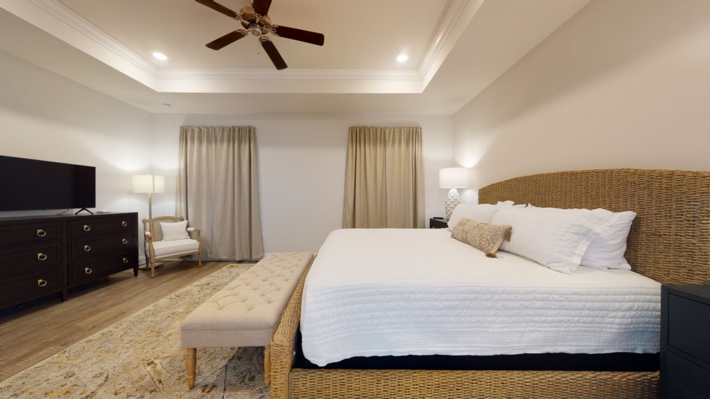 Beautiful, modern primary bed with tray ceiling, ceiling fan, and large flat screen television 
