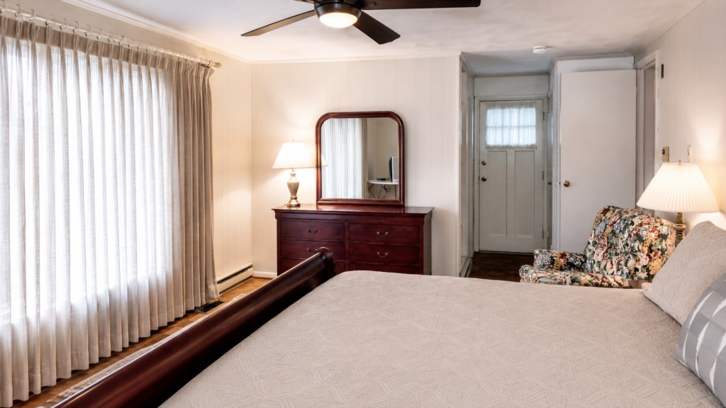 Spacious primary bedroom with large windows, dresser, sitting chair and ceiling fan.